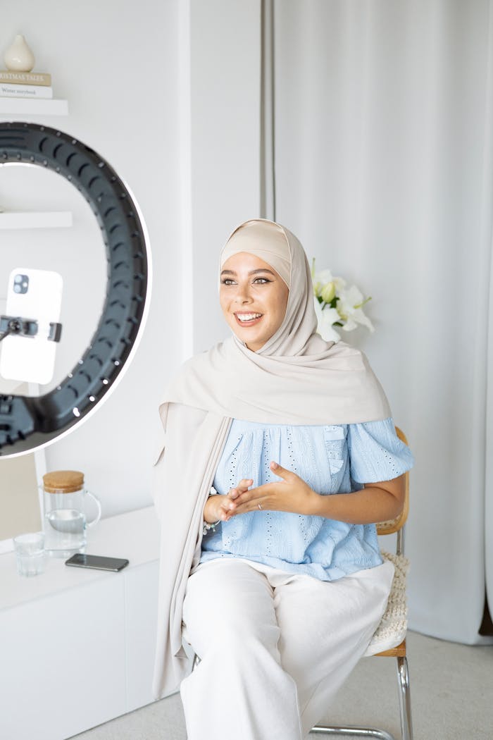 About A Muslim woman wearing a hijab vlogging indoors using a smartphone and ring light.